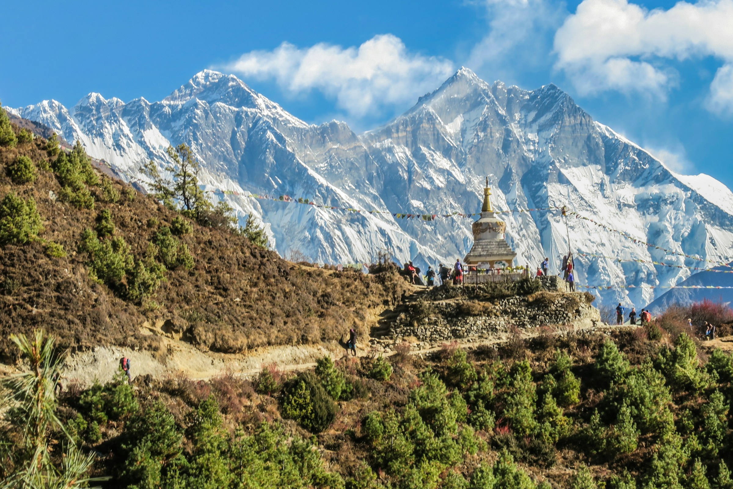 A line of trekkers on a high mountain trail dwarfed by a snow peak behind them