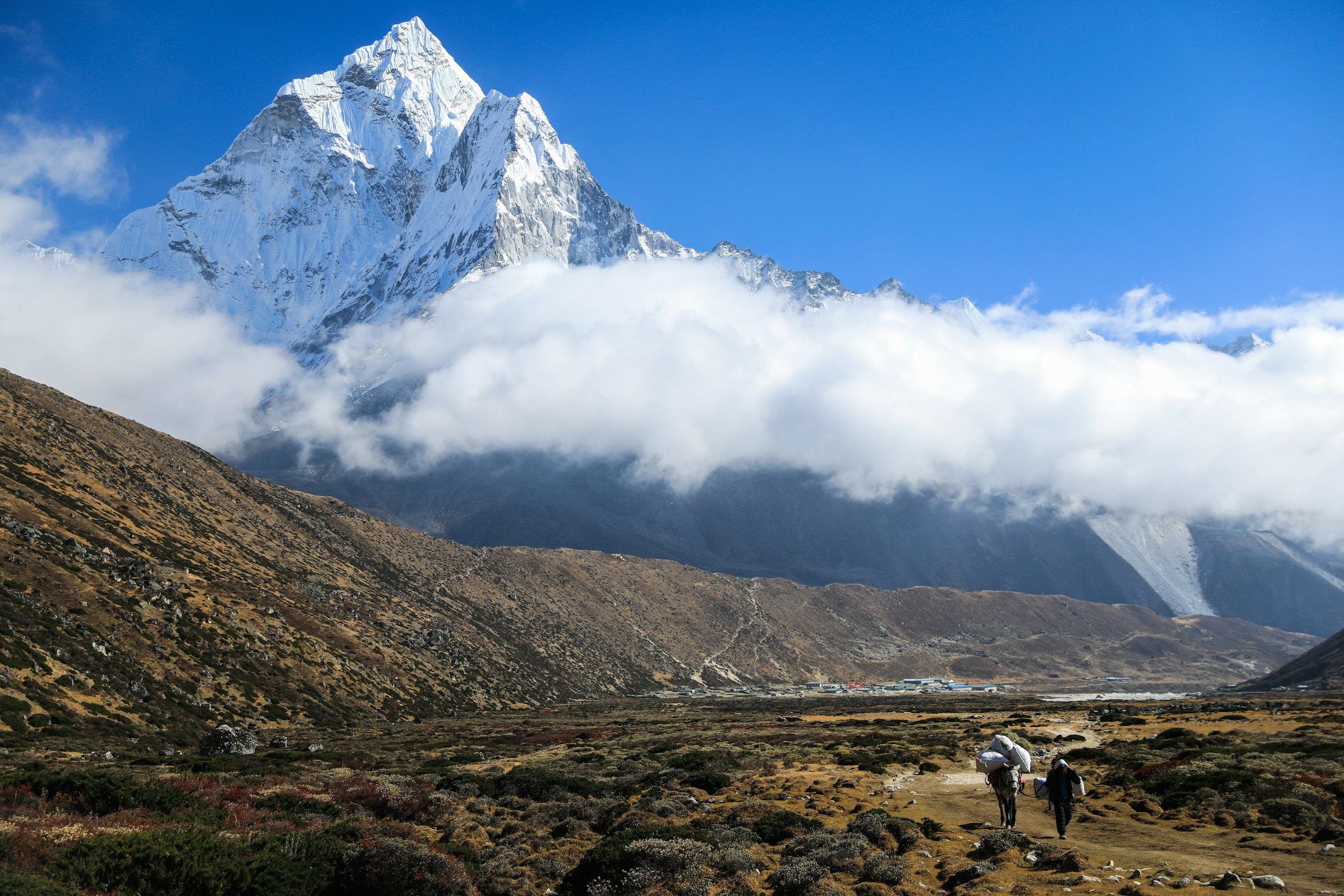 Trekkers on a dirt trail beneath low cloud with Ama Dablam behind them, guided down from Base Camp